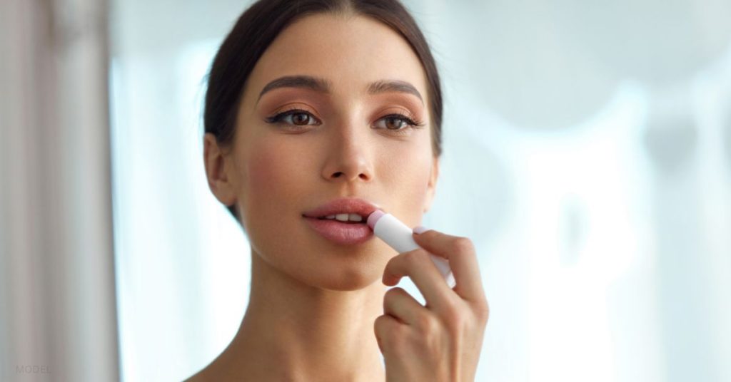 Pretty woman looking in a mirror while applying lip balm to her lips. (model)
