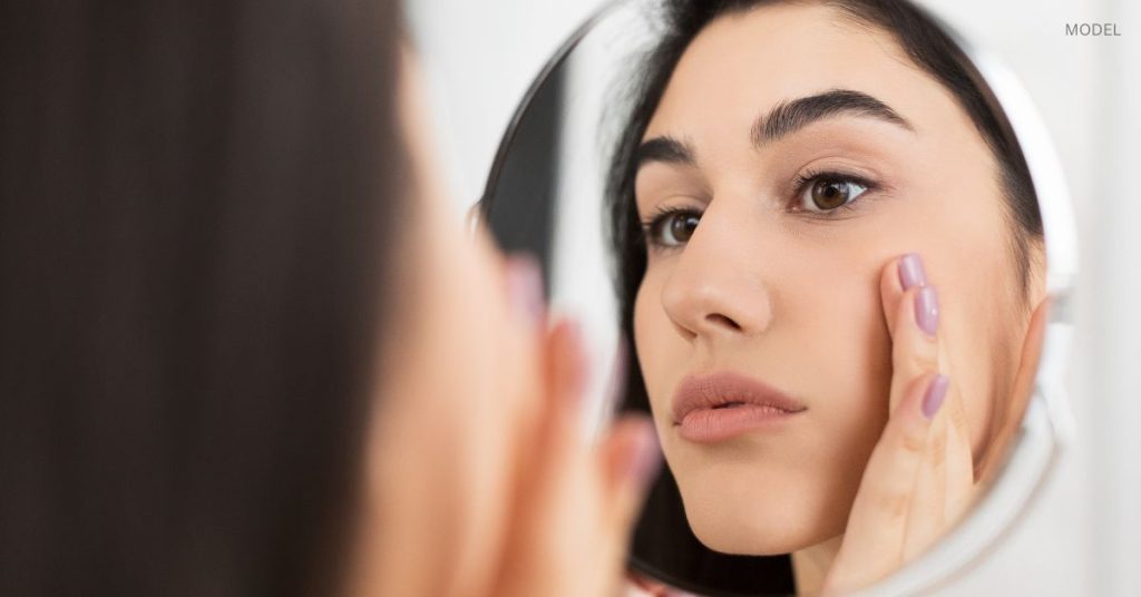 Woman with clear, youthful, beautiful skin (model) looking in the mirror, examining her face.